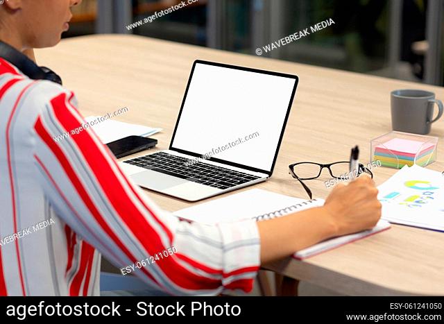 Midsection of biracial female advisor writing in diary by blank screen laptop with copy space
