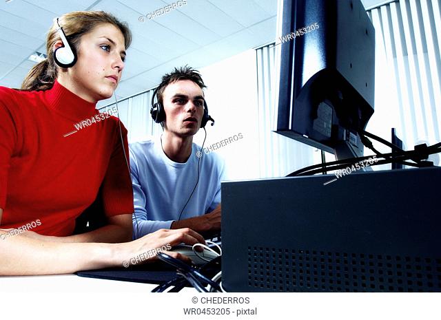 Teenage girl wearing headphones using a computer with a teenage boy sitting beside her