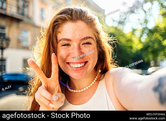 Smiling redhead woman showing peace gesture while taking selfie