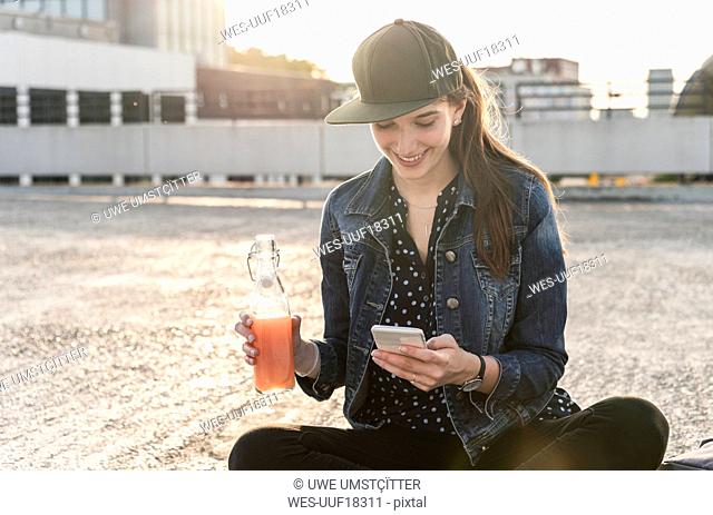 Smiling young woman with drink and cell phone sitting on parking deck at sunset