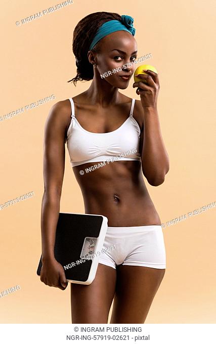 Portrait of attractive african smiling woman eating green apple on beige studio background, looking at camera
