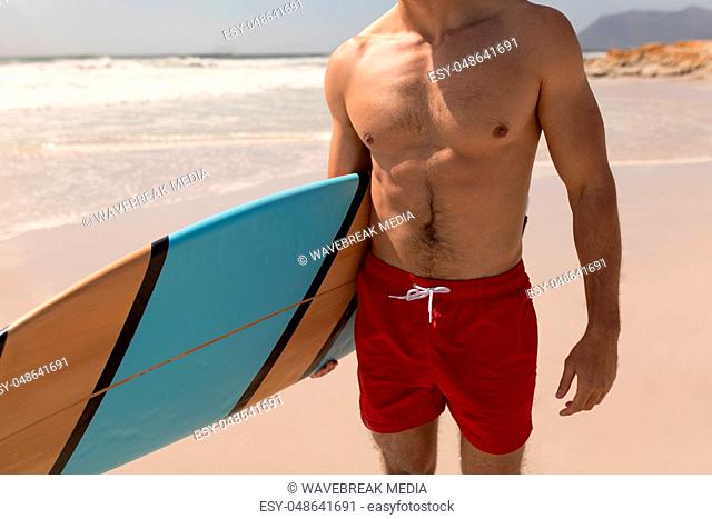 Shirtless young male surfer with surfboard standing on beach