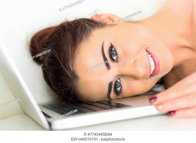 Young smiling woman lying her head on a laptop on a white couch
