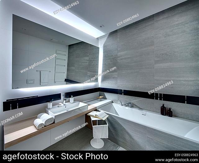 Contemporary bathroom tiled with the white and gray tiles. There is a white sink with accessories, big mirror, bath, towel holder, towel radiator