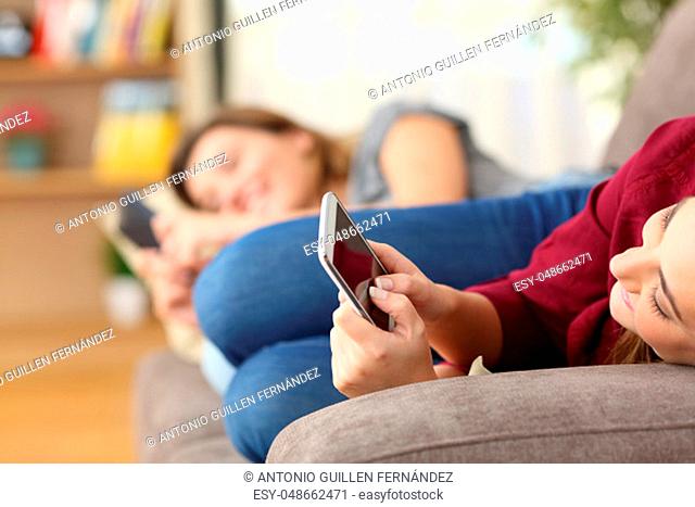 Two roommates using everyone their smart phones lying on a sofa in the living room at home with a homey background