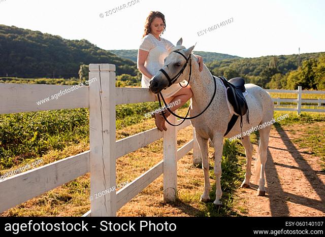 The young woman in a white dress is sitting on the fence and stroking a white horse on a ranch
