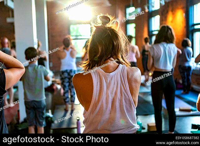 A young Caucasian girl with blonde hair tied up is seen from behind during 108 rounds of surya namaskar sun salutation. Blurred people are seen in background
