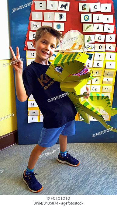 little blond boy with a dino-shaped First-Day-of School-cornet at first day at school, infront of a wall with letters and symbol, Germany