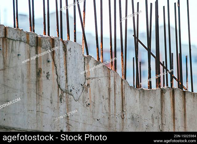 A dilapidated gray concrete wall with rusty rebar protruding from the top of it against a blurred background of an apartment building