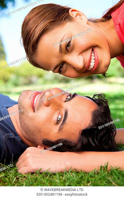 Smiling woman looking towards the side while her head is above her smiling friends head as he is lying on the grass