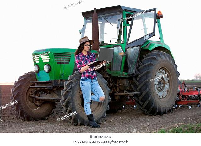 Young country woman with hat holding laptop beside tractor in field