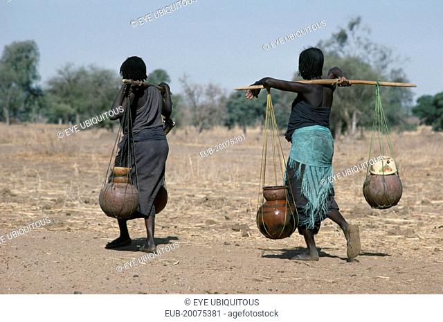 Chadian refugee women carrying water vessels hanging from pole across their shoulders from well in settlement near El Geneina
