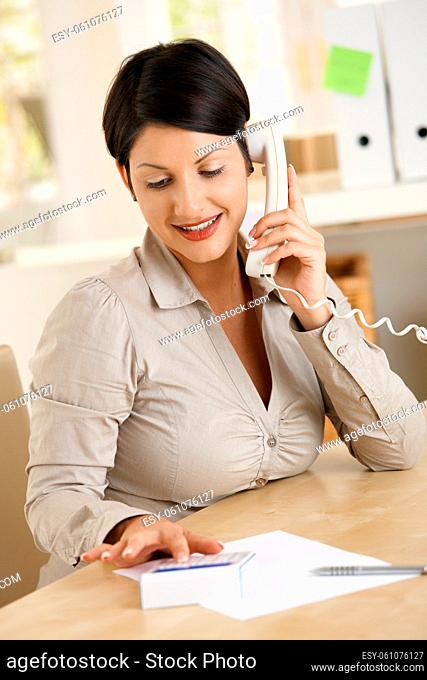 Young businesswoman working at home, talking on phone, using calculator