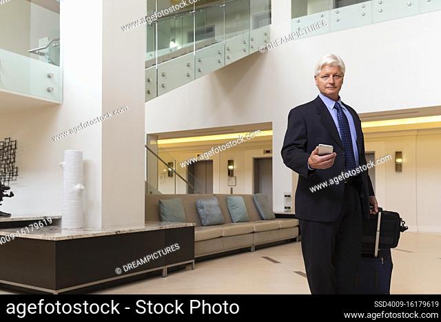 Mature Caucasian man holding cell phone walking through hotel lobby with luggage, looking at camera