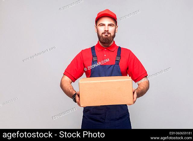 Portrait of young smiling logistic delivery man with beard in blue uniform and red t-shirt standing and holding the cardboard box on grey background