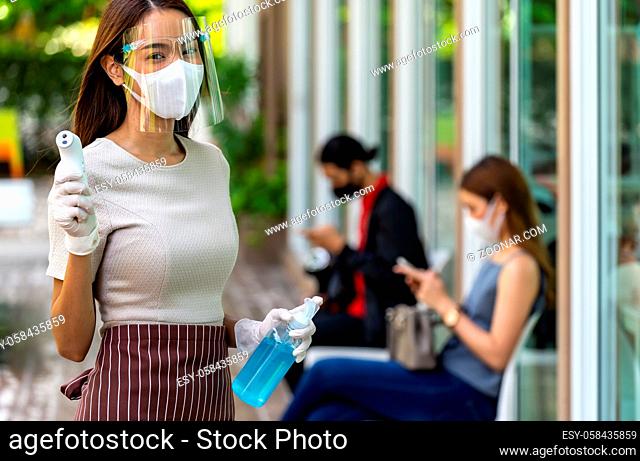 Portrait attractive asian waitress wear face mask and shield hold thermometer and alcohol gel sanitizer with background of social distancing customer waiting in...