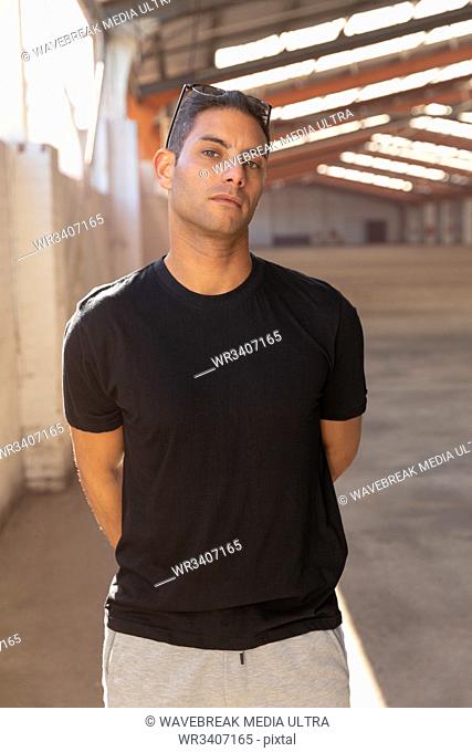 Front view close up of a young Caucasian man wearing a black t shirt and sunglasses on his head, looking to camera with his hands behind his back