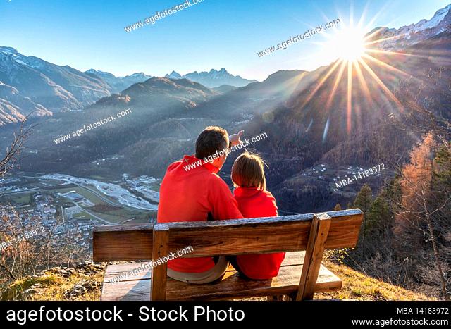 Two people (father and daughter) sitting on a bench in autumn looking at sunset, Agordo, Belluno province, Veneto, Italy