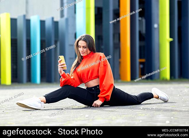 Portrait of beautiful brunette using smart phone while doing splits in front of colorful building