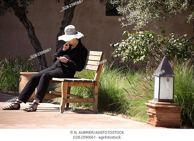 Senior woman with sun hat, sitting on a public wooden bench, reading her tablet, in a garden, Marrakech, Morocco