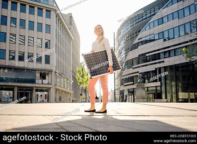 Smiling beautiful female entrepreneur holding solar panel while standing on footpath in financial district