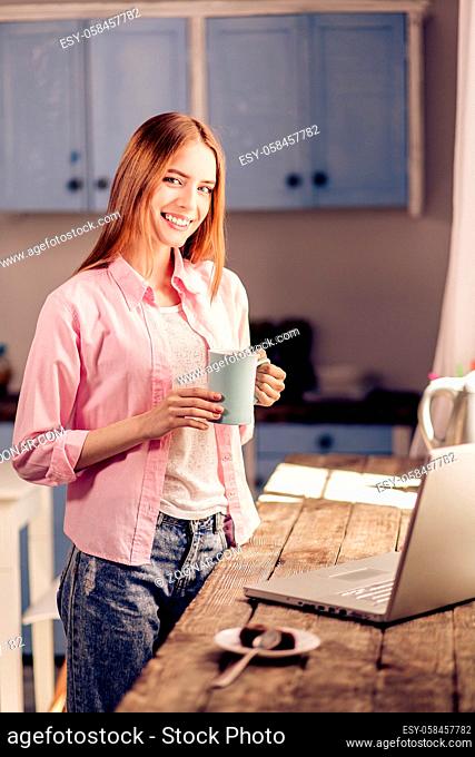 Girl in casual clothes holding cup, standing at home in the kitchen. Pretty young female at home interior close to wooden desk with laptop on it