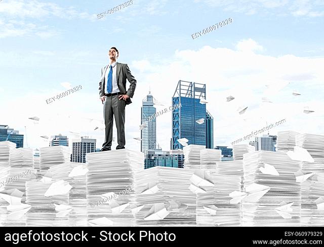 Confident businessman in suit standing on pile of documents among flying paper planes with cityscape on background. Mixed media