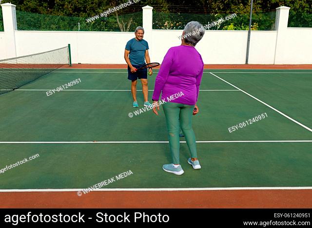 Rear view of biracial senior woman playing tennis with senior man in tennis court during sunny day