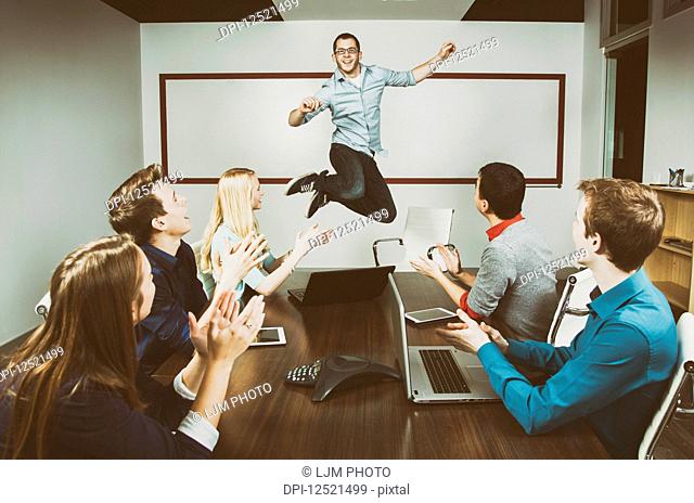 Young millennial business professional jumping to celebrate an accomplishment with his co-workers working in a conference room during a presentation and one of...