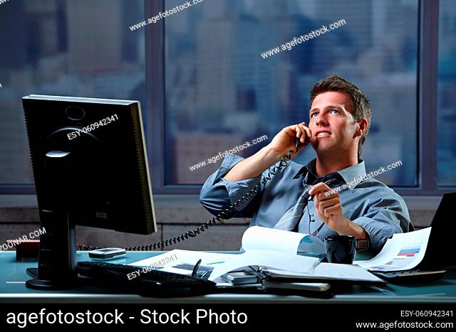 Mid-adult successful smiling businessman calling on landline listening to conversation sitting at office desk