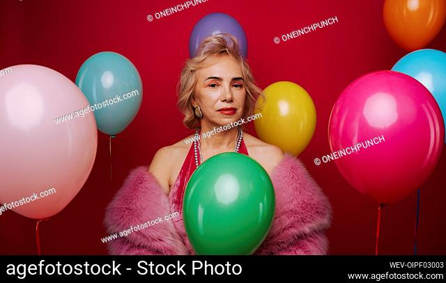 Confident senior woman amidst multi colored balloons against red background