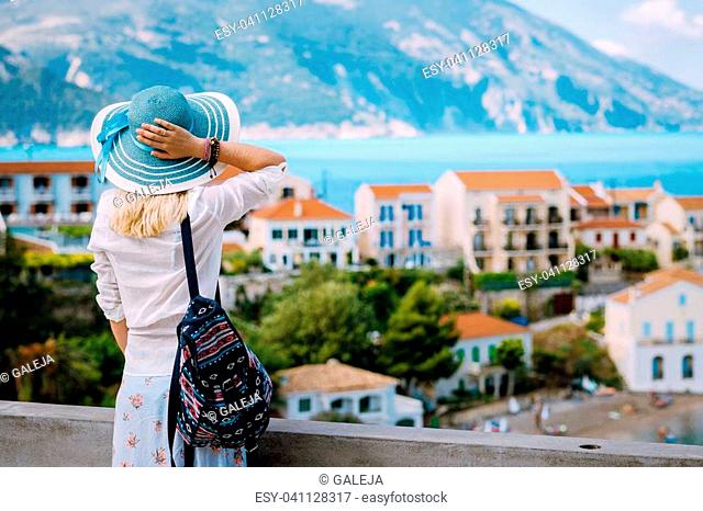 Tourist woman admiring view of colorful tranquil village Assos on morning. Young stylish female model wearing blue sunhat and white clothes enjoying summer...