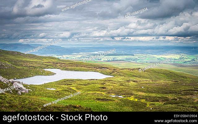 Green fields or rolling hills with small lake at footstep of Cuilcagh Mountain, dramatick stormy sky in background, Northern Ireland