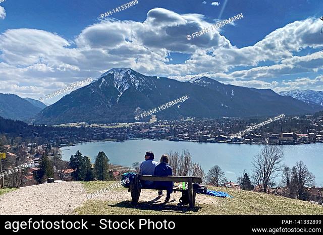 Couple takes a rest on a bench. Hikers on the Hoehenweg over the Tegernsee with a view of Rottach Egern on April 1st, 2021