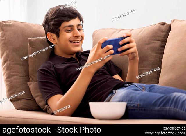 A TEENAGE BOY HAPPILY USING MOBILE PHONE WHILE RESTING ON SOFA