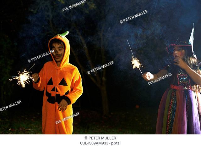 Brother and sister wearing halloween costumes holding sparklers in garden at night