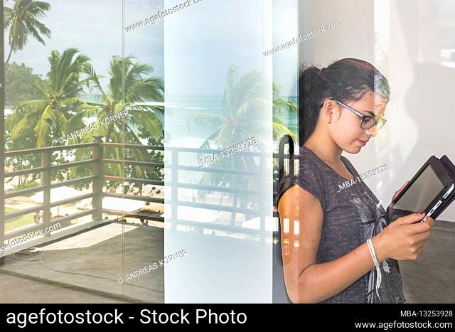 young woman stands leaning against patio door with her tablet on vacation on a palm beach
