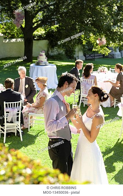Mid adult bride and groom in garden toasting among wedding guests