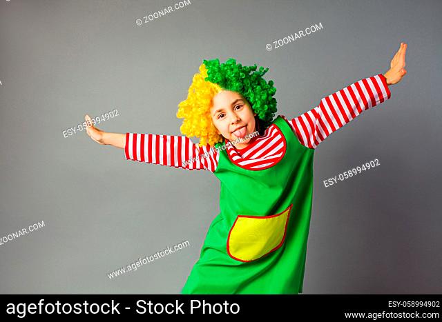 The little girl in a clown uniform stands on a gray background. The child is posing with his tongue outstretched and spreads her arms