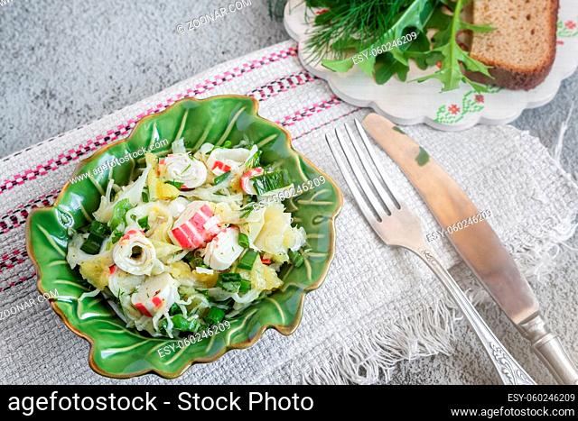On the table in a ceramic plate vegetable salad of cabbage, pepper, green onions, crab sticks. Next to the spices and bread