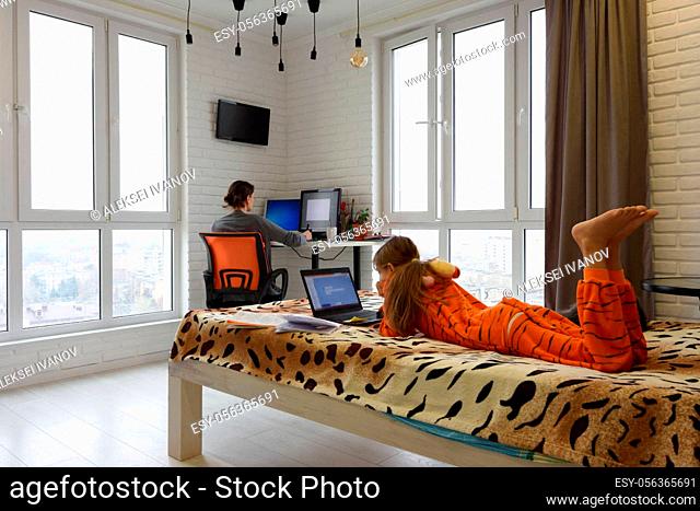 The family is at home, the child lies on the bed and studies in a laptop, the mother in the background works remotely in the computer