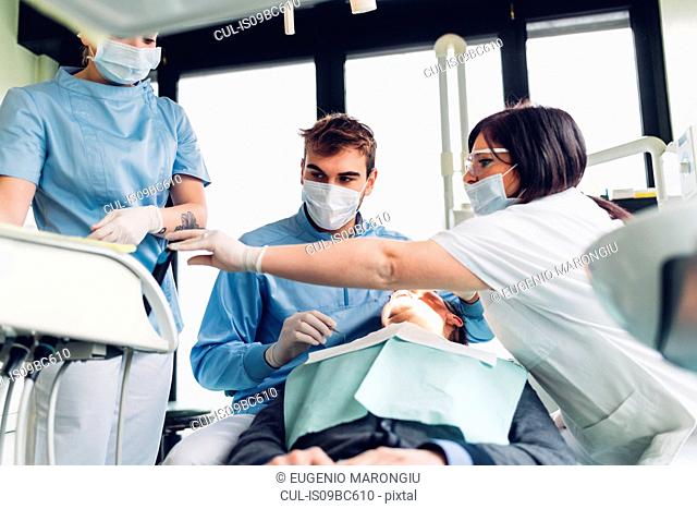 Dentist looking into male patient's mouth, dental nurses preparing equipment