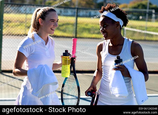 Smiling biracial female tennis players with bottles and towels talking during break at court