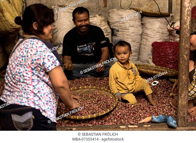 A toddler sitting in the market stall of Kutacane between many onions in his family's market stall