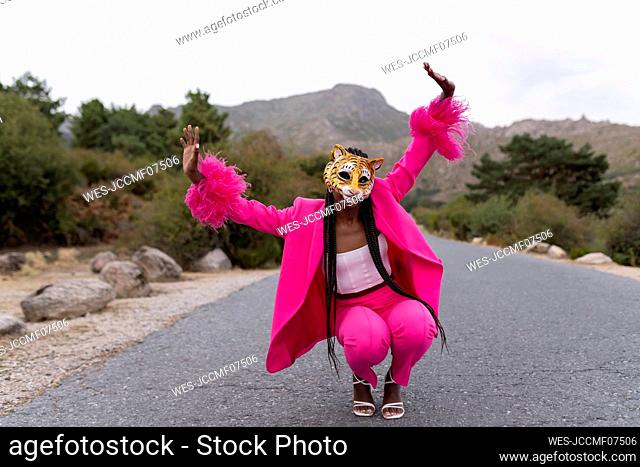 Young woman wearing tiger mask crouching on road with arms raised