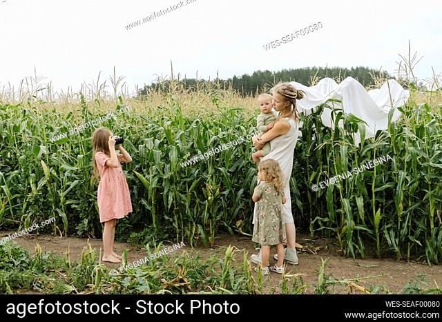 Girl photographing mother and sibling standing near corn field