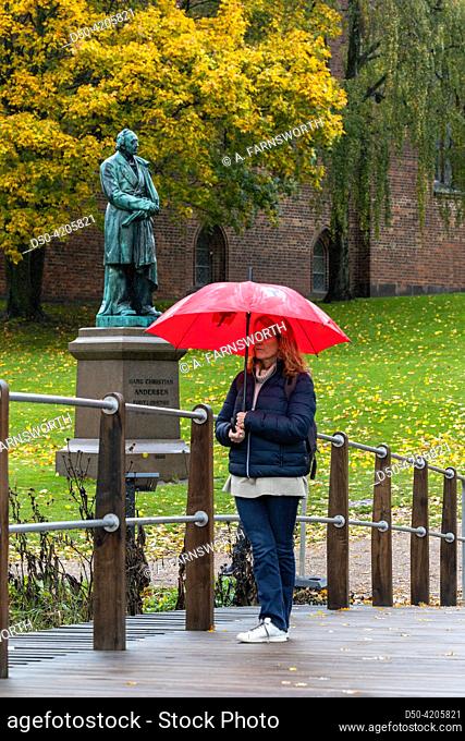 Odense, Denmark A woman with a red umbrella stand near statue of HC Andersen in a park