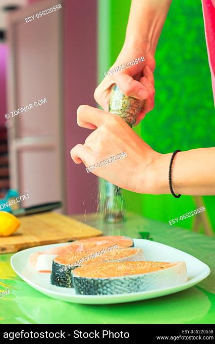 women's hands sprinkle from the hand mill with spices two raw steak from salmon lying on a white plate in a home kitchen in green tones