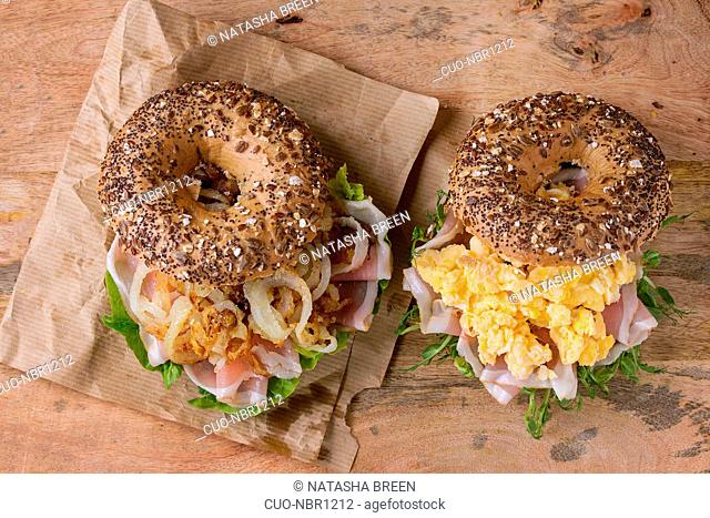 Two Whole Grain bagels with fried onion, scrambled eggs, green salad and prosciutto ham on paper over wooden textured background. Top view