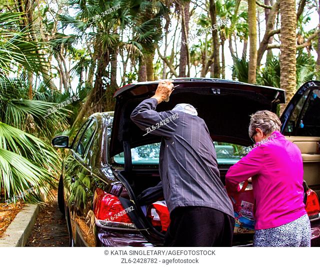 Older caucasian couple checking the trunk of their car in the middle of a palm trees parking in the nature
